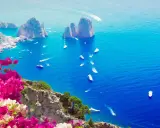 Un giorno a Capri: scogliere rocciose, mare blu intenso e barche che solcano le onde, con bouganville rosa in primo piano.