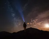 Una persona con una torcia illumina il cielo stellato su una montagna di notte. La Via Lattea è visibile.