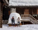Coppia con racchette da neve a Riale Val Formazza, con un drone in volo sopra un tetto innevato.