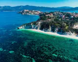 Vista aerea di una spiaggia bianca con acque cristalline e un borgo storico su una collina, parte dell'arcipelago toscano isole visitabili.