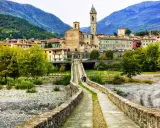 Ponte Gobbo e borghi vicino a Bobbio, con montagne sullo sfondo. Due persone passeggiano sul ponte antico.