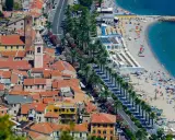Aerial view of a coastal town with red-tiled roofs, a beach with umbrellas, and palm trees lining the road.