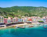Colorate case affacciate sul mare cristallino, con barche ormeggiate e la collina verde sullo sfondo. Un'immagine che evoca il fascino del traghetto per l'Isola del Giglio.