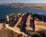 Borgo medievale fortificato domina la campagna toscana lungo il mare, con un castello imponente e case dai tetti rossi affacciate su un golfo con barche a vela.