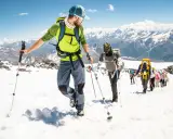 Un gruppo di escursionisti sta camminando sulla neve in montagna, con bastoncini da trekking e zaini, sotto un cielo azzurro.