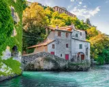 Antichi borghi medievali lombardia da visitare: un ponte di pietra collega case storiche sull'acqua cristallina, con edera rampicante e vegetazione lussureggiante.