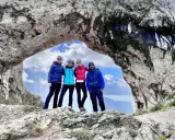 Quattro amici si godono una giornata di camminare in montagna, posando sotto un arco naturale di roccia con un cielo azzurro e nuvoloso.