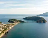 Panoramica di un'isoletta tra Napoli e Ischia, con un borgo costiero, un porto turistico e un ponte che collega a un'altra isola verdeggiante.