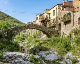 Antico ponte in pietra collega i borghi dell'entroterra ligure ponente, con case colorate e vegetazione rigogliosa che scende verso il torrente.