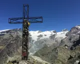 Croce metallica e di pietra in cima a una montagna, con un ghiacciaio e vette innevate sullo sfondo. Il **palon di Resy** domina il panorama.