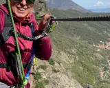 Donna sorridente si aggrappa a un cavo d'acciaio, godendosi un'avventura sulle ferrate Val di Susa con un panorama mozzafiato.