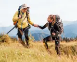 Coppia di escursionisti si aiutano a vicenda su un sentiero di montagna, con zaini e bastoncini da trekking.