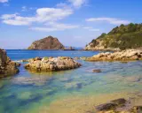 Acque cristalline e rocce scolpite dal mare a Porto Ercole, con montagne verdi sullo sfondo e un cielo azzurro punteggiato di nuvole.