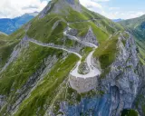Un tortuoso cammino del sale si snoda tra montagne verdi, con ciclisti che affrontano tornanti mozzafiato su un sentiero panoramico.