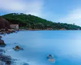 Una casa di pietra sulla spiaggia di Bengodi, con il mare calmo e una collina verdeggiante sullo sfondo.