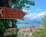 Segnale arancione indica "Sentiero del Viandante" con vista su un lago e montagne.