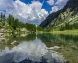 Specchio d'acqua cristallino tra i **laghi della Valle d'Aosta**, circondato da pini e rocce, con montagne imponenti e cielo azzurro con nuvole bianche.