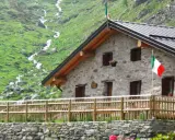Rifugio in pietra con bandiera italiana, circondato da verdi pendii e un ruscello. Un'oasi accogliente tra i rifugi Val Camonica facili da raggiungere.