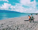 Due ragazzi passeggiano sulla spiaggia della Maremma Toscana, uno con una tavola da surf, mentre il mare azzurro lambisce la riva.