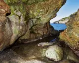 L'entrata di una grotta marina, la cala del bove, con rocce muschiose e un piccolo specchio d'acqua che riflette il blu del mare e il cielo.