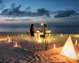 Una coppia al mare di sera, seduta a tavola per una cena romantica illuminata da lanterne e luci soffuse sulla sabbia.