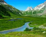 Val Ferret, un paradiso per passeggiate facili. Un fiume serpeggia tra prati verdi e montagne innevate sotto un cielo azzurro.