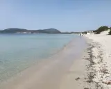 Spiaggia della Fertilia con acque cristalline, sabbia bianca e un edificio in lontananza. Due persone passeggiano lungo la riva.