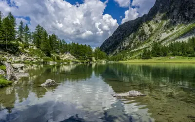 Specchio d'acqua cristallino tra i **laghi della Valle d'Aosta**, circondato da pini e rocce, con montagne imponenti e cielo azzurro con nuvole bianche.