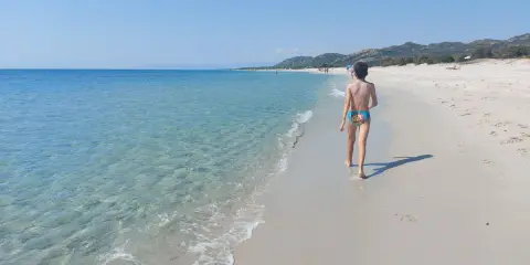 Un bambino cammina su una spiaggia di sabbia bianca, verso le spiagge più belle della Sardegna est. Acqua cristallina e cielo azzurro.
