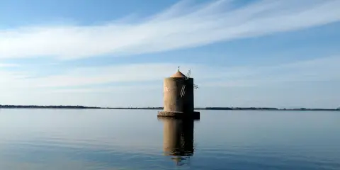Mulino solitario nella laguna di Orbetello, con cielo azzurro e nuvole sottili.