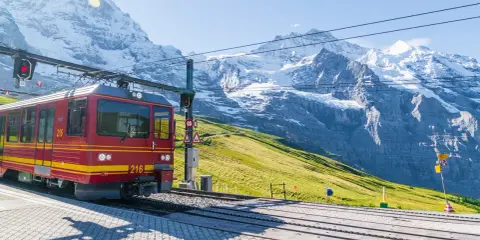 Treno rosso e giallo in stazione, con imponenti montagne innevate sullo sfondo. Un viaggio in treno tra le vette.