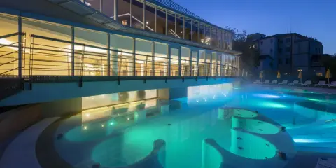 Piscina termale illuminata di notte, con vapori che salgono. Un edificio moderno con ampie vetrate si affaccia sull'acqua turchese, evocando un'atmosfera di relax e benessere, come in antiche terme preistoriche a Padova.