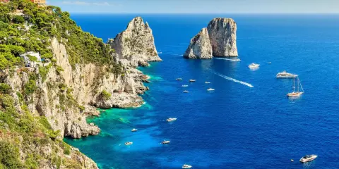 Scogliere verdi e rocciose si affacciano sul blu intenso del golfo di Napoli, punteggiato da barche e yacht. Faraglioni iconici emergono dal mare.