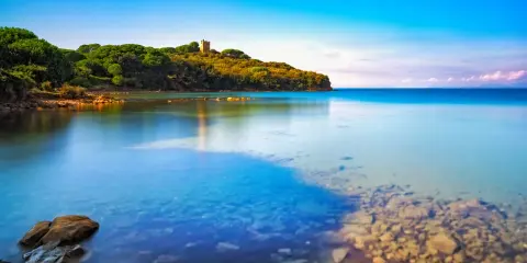 Torre medievale su una collina verdeggiante affacciata sul limpido **golfo di Follonica**. Acque calme e trasparenti riflettono il cielo azzurro.