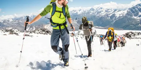 Un gruppo di escursionisti sta camminando sulla neve in montagna, con bastoncini da trekking e zaini, sotto un cielo azzurro.