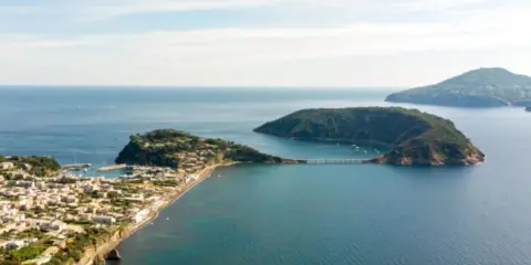 Panoramica di un'isoletta tra Napoli e Ischia, con un borgo costiero, un porto turistico e un ponte che collega a un'altra isola verdeggiante.