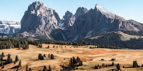 Un paesaggio mozzafiato delle Dolomiti più belle, con cime imponenti, prati dorati e boschi verdi. Piccole baite sparse creano un'atmosfera idilliaca.