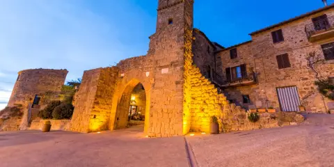 Antica porta e torre medievale illuminata, con case in pietra. Monticchiello cosa vedere: un borgo suggestivo.