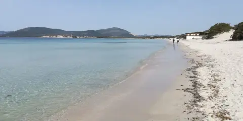 Spiaggia della Fertilia con acque cristalline, sabbia bianca e un edificio in lontananza. Due persone passeggiano lungo la riva.