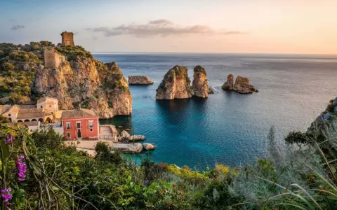 Un borgo con torri antiche e una casa rossa si affaccia su un mare bello Italia, con faraglioni che emergono dalle acque cristalline.