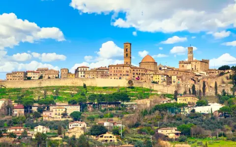 Panoramica di Volterra, dove si trova questo antico borgo toscano, con le sue torri e case arroccate sulla collina sotto un cielo azzurro con nuvole bianche.