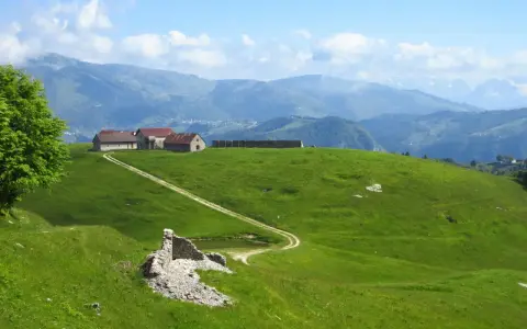 Un sentiero delle malghe serpeggia tra verdi colline, conduce a rustici edifici e si perde tra le montagne nebbiose.