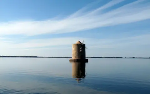 Mulino solitario nella laguna di Orbetello, con cielo azzurro e nuvole sottili.