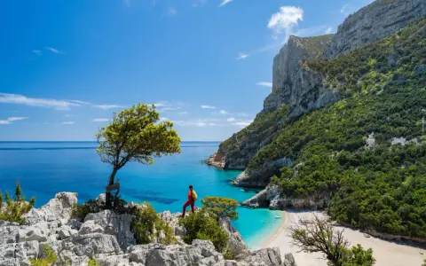 Escursionista ammira una baia cristallina con una spiaggia bianca, incastonata tra imponenti scogliere verdi. Un paesaggio mozzafiato che ricorda la bellezza della montagna centro Italia.