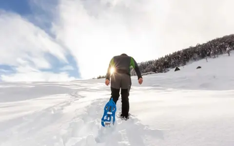 Uomo con ciaspole cammina su sentiero innevato, ideale per chi cerca dove ciaspolare in Piemonte. Sole splendente tra le nuvole.