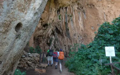 Escursionisti esplorano un sentiero nella Riserva dello Zingaro, avvicinandosi a un'imponente parete rocciosa con vegetazione rigogliosa.