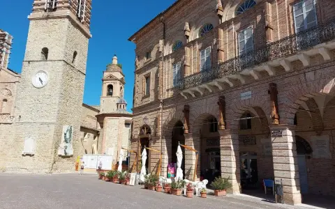 Piazza a San Ginesio, Marche, con il campanile in pietra e l'edificio in mattoni rossi con portici.