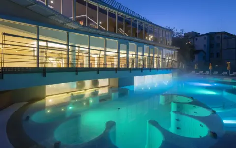 Piscina termale illuminata di notte, con vapori che salgono. Un edificio moderno con ampie vetrate si affaccia sull'acqua turchese, evocando un'atmosfera di relax e benessere, come in antiche terme preistoriche a Padova.