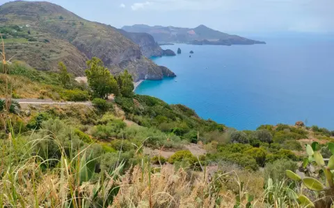 Panoramica mozzafiato per capire come visitare le Isole Eolie: costa verdeggiante, mare blu intenso e isole vulcaniche all'orizzonte.