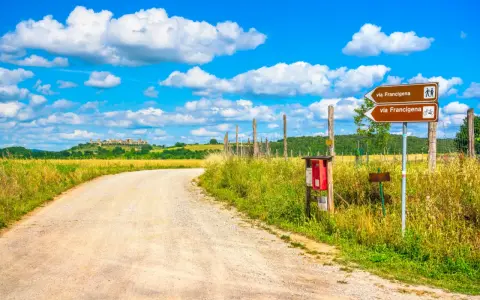 Segnaletica per la Via Francigena, un antico cammino. La strada sterrata conduce verso un borgo collinare sotto un cielo azzurro con nuvole bianche.